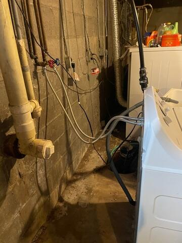 A dark utility corner of a concrete block basement next to water pipes and a white washing machine in Wrenshall, MN before waterproofing.