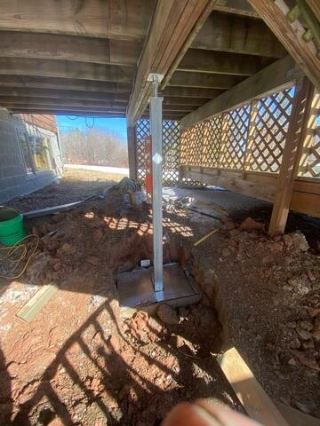active-helical-pier-foundation-repair-grand-marais-mn An active structural foundation repair site underneath a deck in Grand Marais, MN, showing a deep excavated pit and a newly installed galvanized steel support pier column.