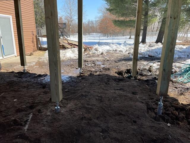 An under-deck view looking out toward a snowy yard showing several high-capacity galvanized steel helical pier columns supporting timber posts in Bethel, MN.