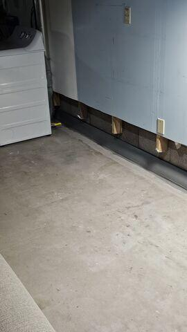 A clean concrete basement floor showing a newly installed grey DryTrak baseboard drainage system running along the base of a foundation wall next to a white washing machine appliance in Bayfield, WI.
