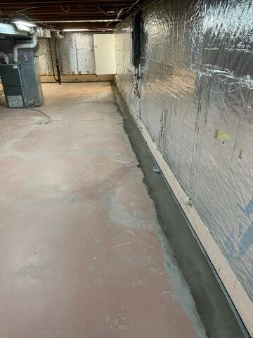 A clean basement floor showing a freshly poured concrete trench patch alongside a wall lined with silver reflective insulation sheets during a waterproofing project in Cornell, WI.