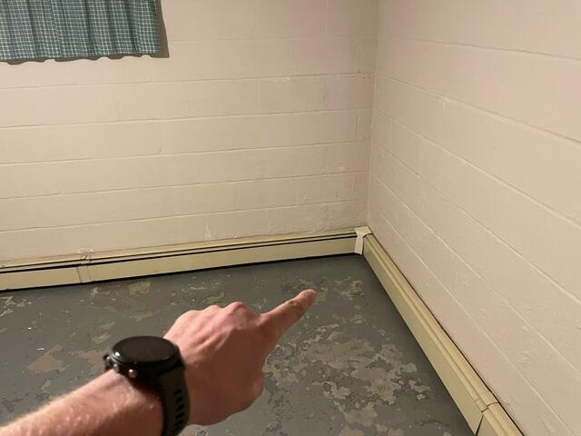 A hand pointing to a corner wall junction with peeling concrete floor paint and moisture damage in an Esko, MN basement before repair.