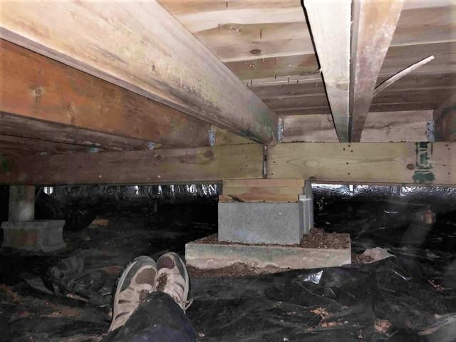 A low-clearance crawl space with a black plastic vapor barrier on the dirt floor, showing heavy timber joists supported by a concrete block pier column in Couderay, WI during a foundation inspection.