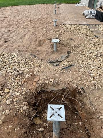 A row of heavy-duty steel helical pier brackets and galvanized shafts installed deep inside excavated ground trenches in a yard in Shell Lake, WI.