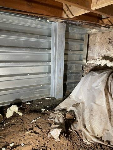 A close-up view of an installed EverBrace corrugated steel wall panel and a vertical steel I-beam brace next to a crawl space dirt floor in Frederic, WI.