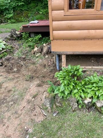 An exterior view of a log-sided cabin built on a grassy hillside with excavated dirt beds and a wooden deck in Arkansaw, WI.