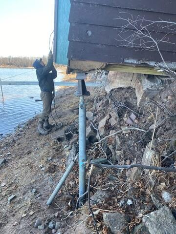A technician working near an installed galvanized steel helical pier supporting the corner baseline of a brown wooden boathouse on the shore of Sturgeon Lake, MN.