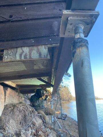 A close-up view of a technician wearing a welding helmet creating sparks while welding a heavy steel bracket to a helical pier foundation column under a boathouse in Sturgeon Lake, MN.